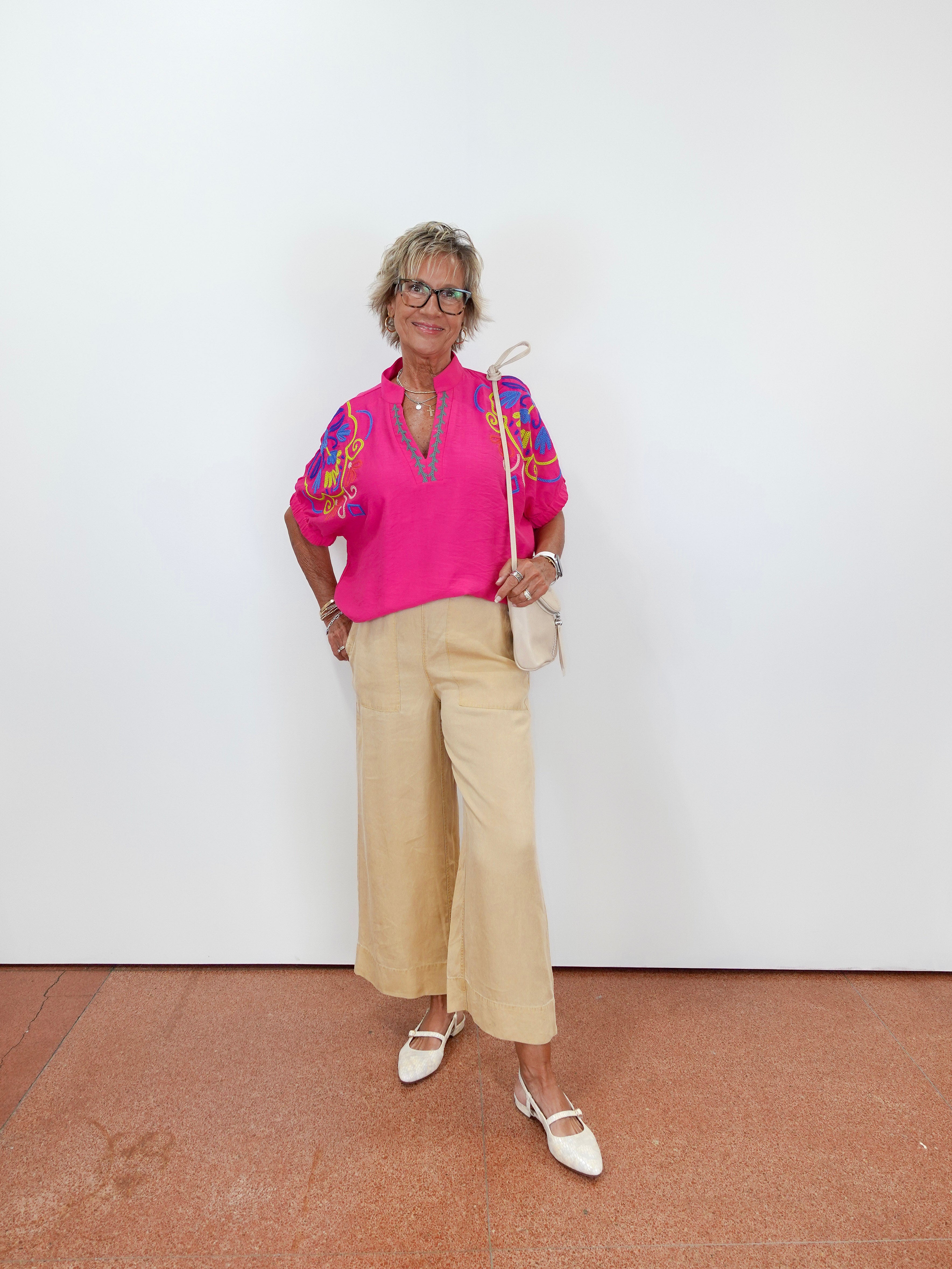 Woman wearing a bright pink top with colorful patterns and beige pants against a white wall.