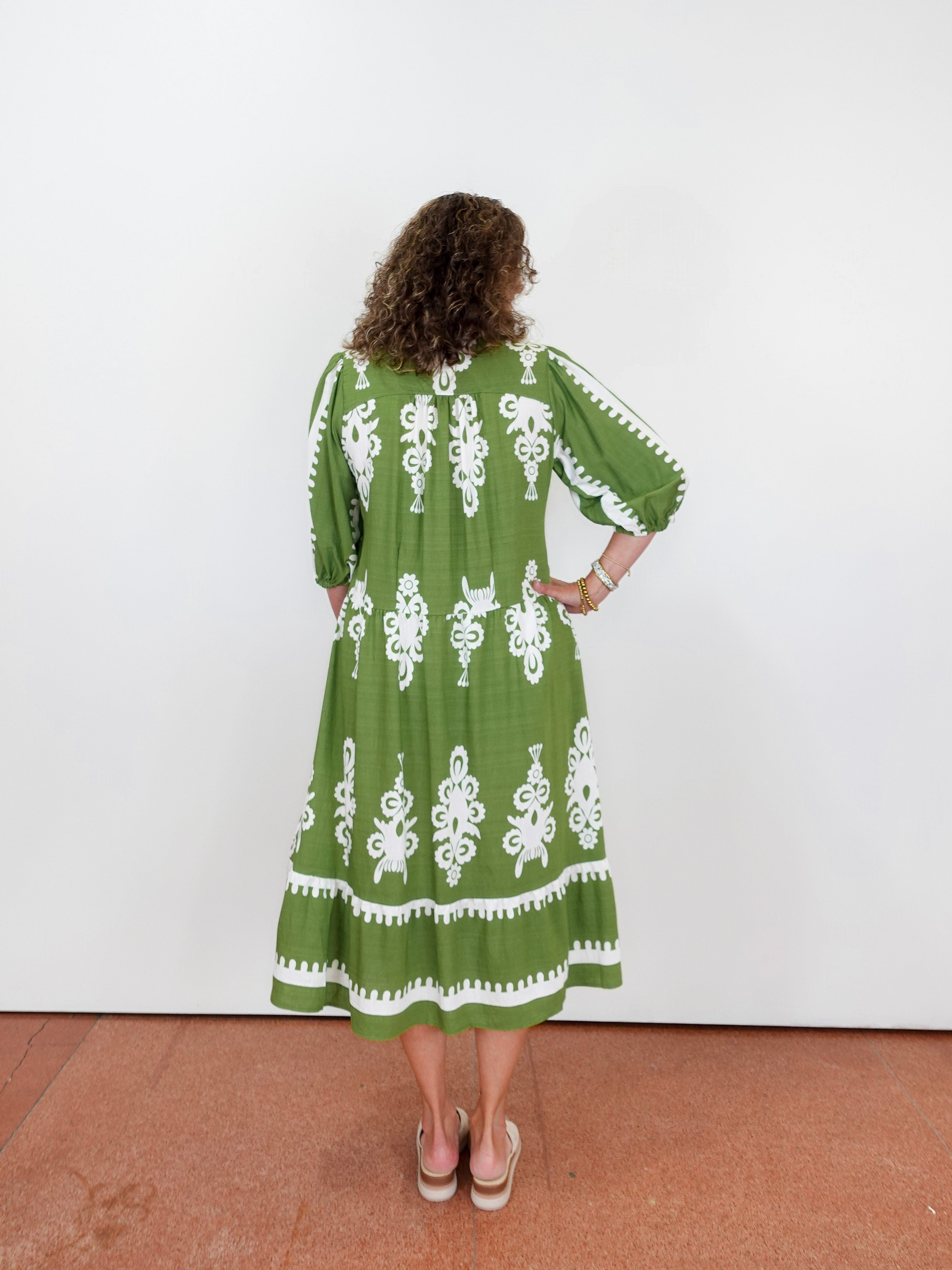 Woman wearing a green dress with white patterns standing against a white wall.