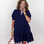Woman wearing a navy blue dress with a white collar, standing against a plain background.