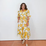 Woman wearing a yellow and white floral dress standing against a white wall.