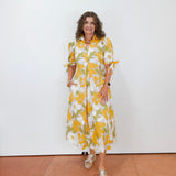Woman wearing a yellow and white floral dress standing against a white wall.