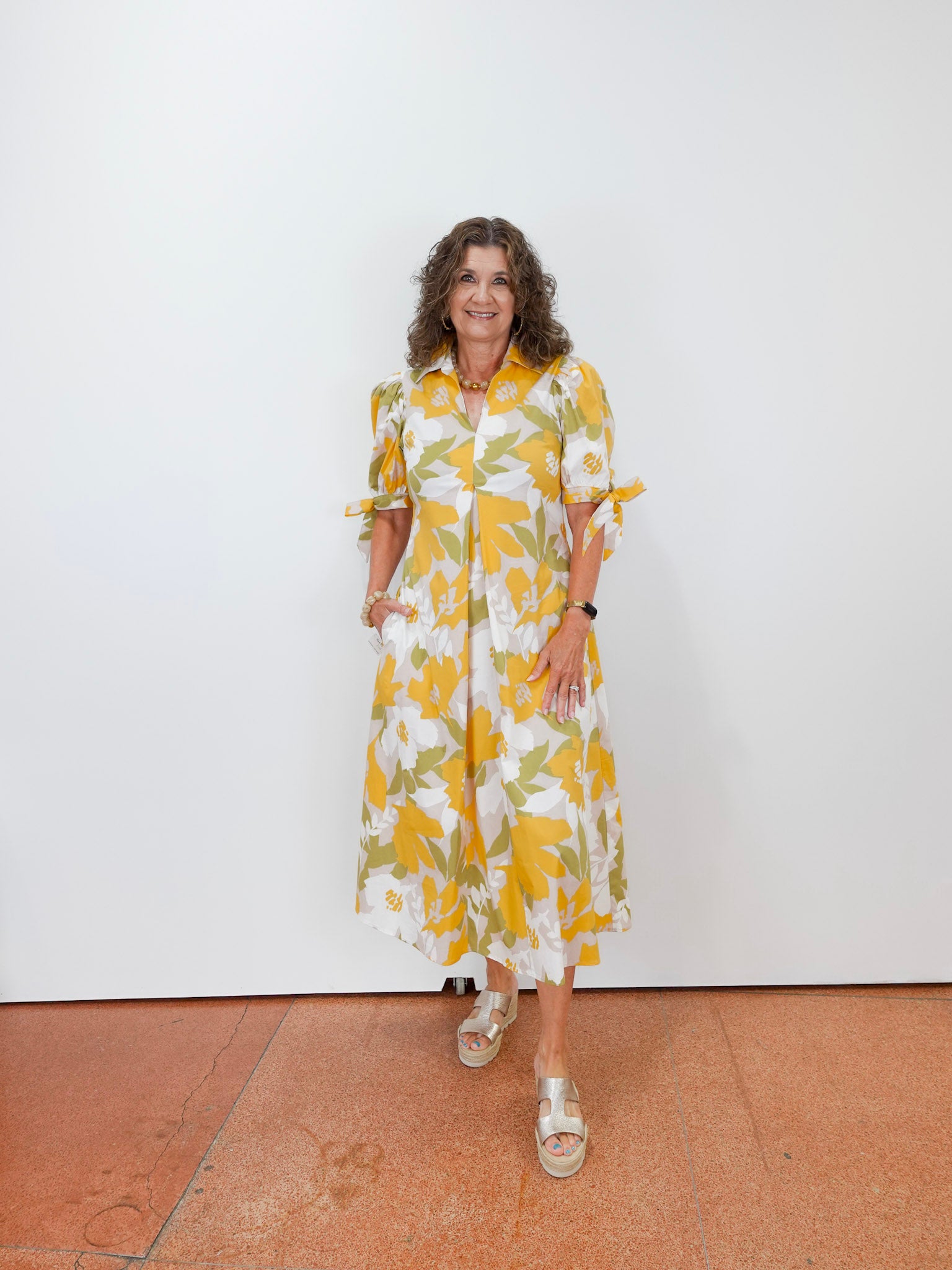 Woman wearing a yellow and white floral dress standing against a white wall.