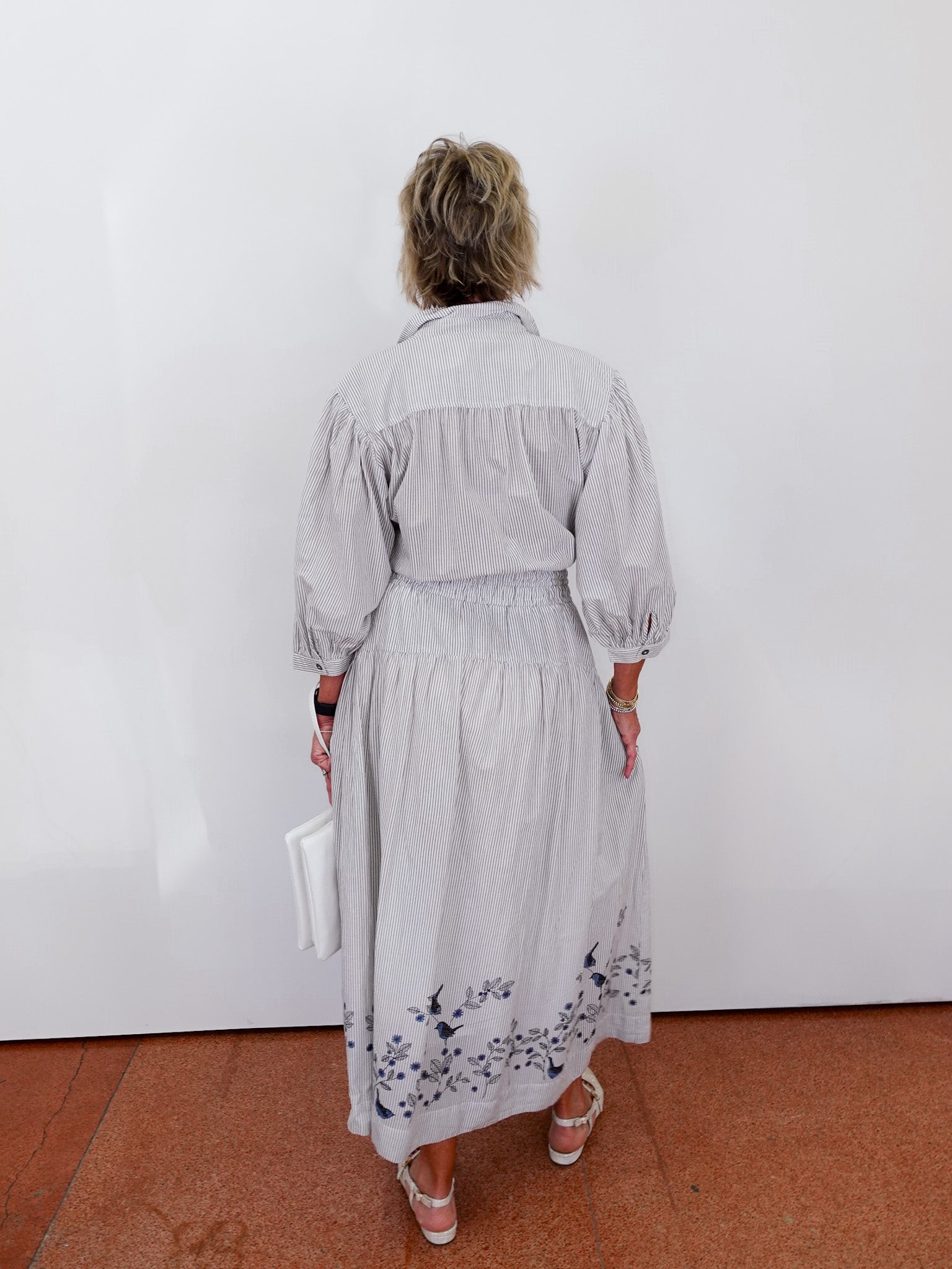Person wearing a light gray dress with floral patterns, standing against a white wall.