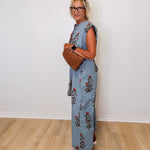 Woman wearing a floral jumpsuit holding a brown leather bag against a white wall.