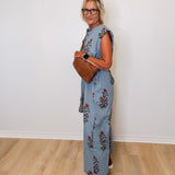 Woman wearing a floral jumpsuit holding a brown leather bag against a white wall.