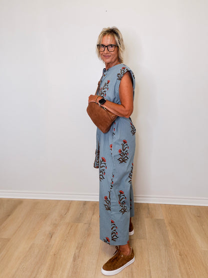 Woman wearing a floral jumpsuit holding a brown leather bag against a white wall.