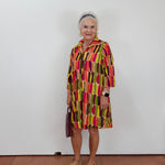 An older woman stands against a plain white wall wearing a colorful abstract-patterned dress with vertical blocks of red, yellow, green, and brown. She is holding a dark handbag and wearing tan slide sandals.