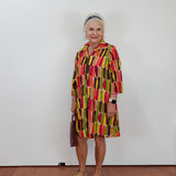 An older woman stands against a plain white wall wearing a colorful abstract-patterned dress with vertical blocks of red, yellow, green, and brown. She is holding a dark handbag and wearing tan slide sandals.