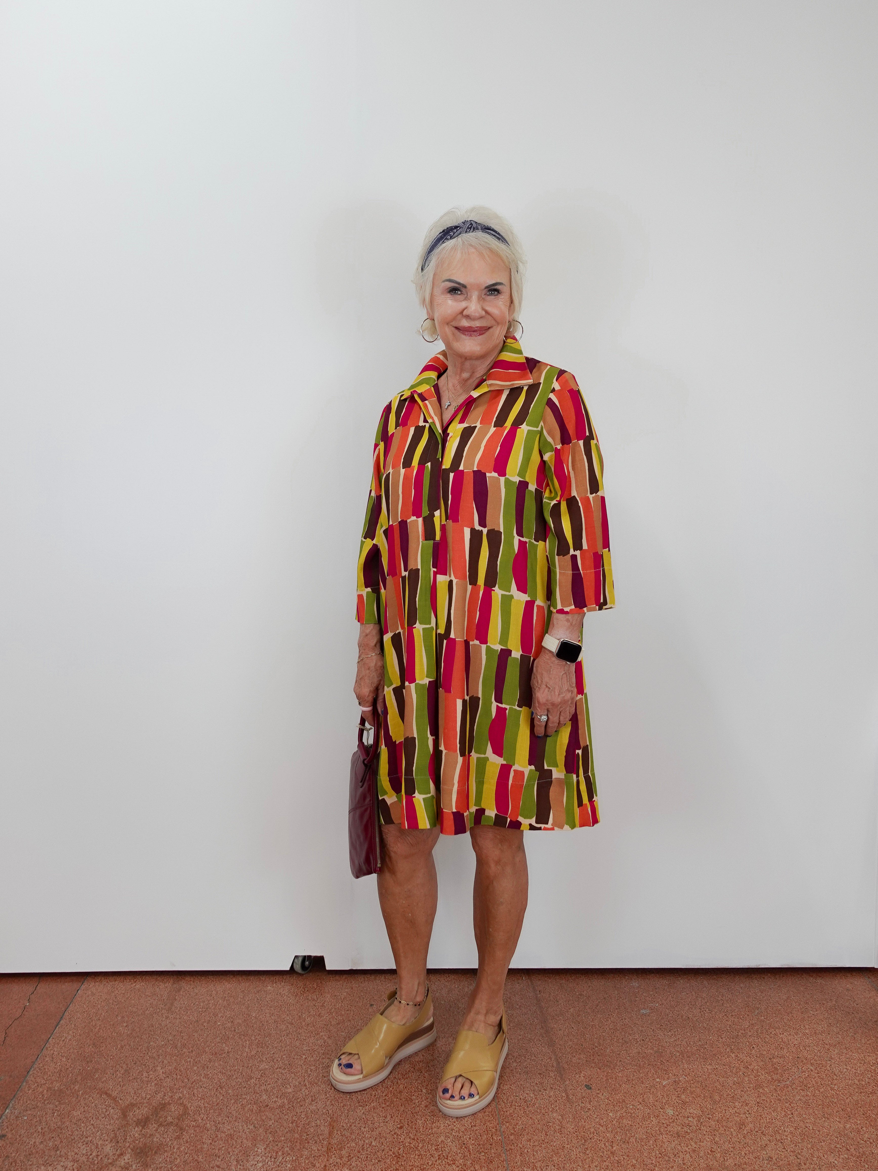 An older woman stands against a plain white wall wearing a colorful abstract-patterned dress with vertical blocks of red, yellow, green, and brown. She is holding a dark handbag and wearing tan slide sandals.