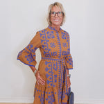 Woman wearing a brown dress with blue patterns against a white wall.