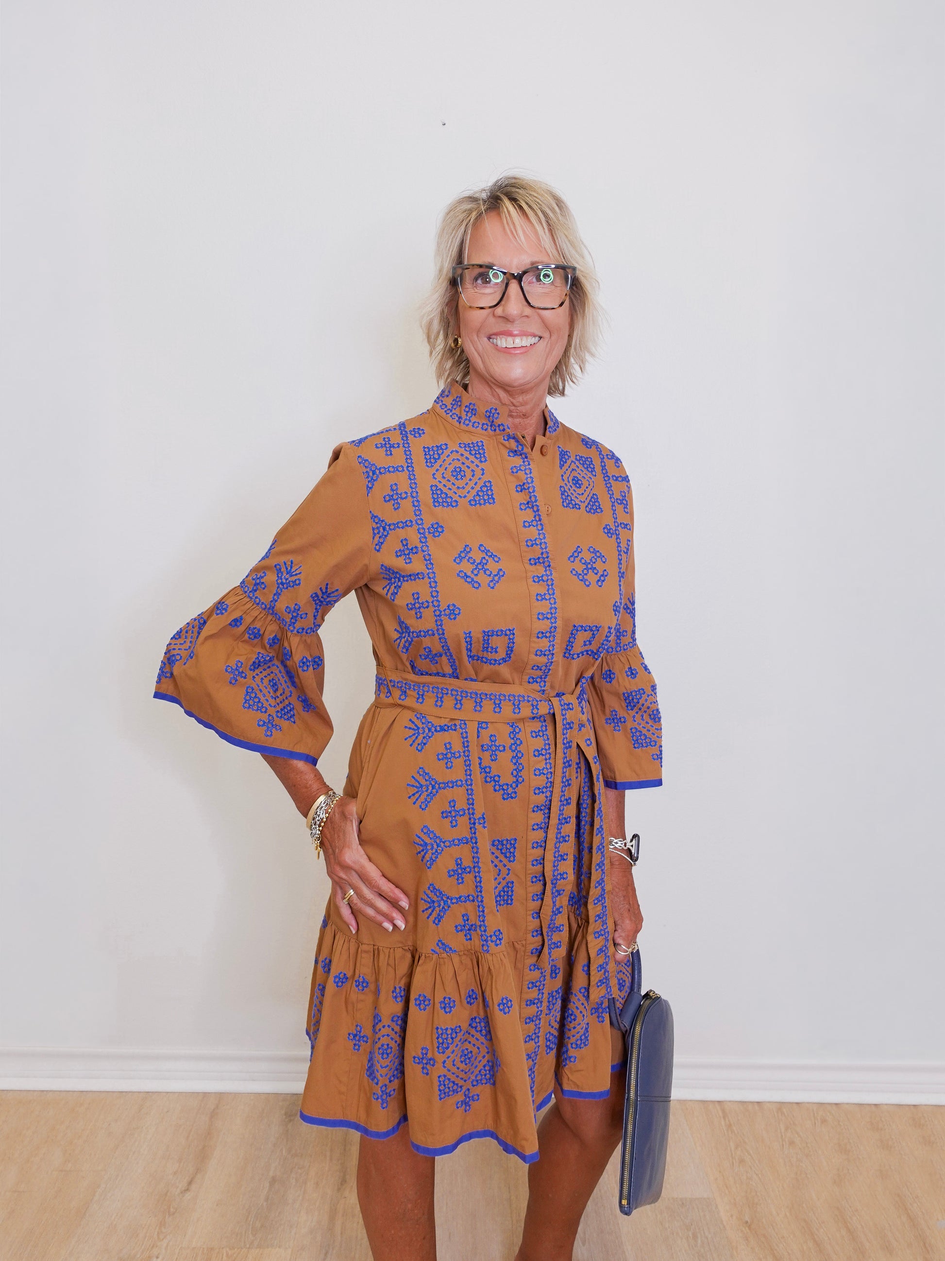 Woman wearing a brown dress with blue patterns against a white wall.