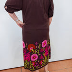 A woman wearing a dark brown off-the-shoulder midi dress with colorful floral embroidery along the hem, paired with beige wedge sandals, shown from the back against a white wall.