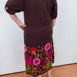 A woman wearing a dark brown off-the-shoulder midi dress with colorful floral embroidery along the hem, paired with beige wedge sandals, shown from the back against a white wall.