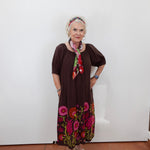 An older woman stands against a plain white wall wearing a dark brown short-sleeve dress with colorful floral embroidery at the hem, a multicolor floral headband, and a matching neck scarf.