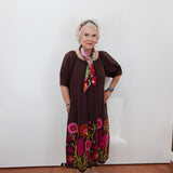 An older woman stands against a plain white wall wearing a dark brown short-sleeve dress with colorful floral embroidery at the hem, a multicolor floral headband, and a matching neck scarf.