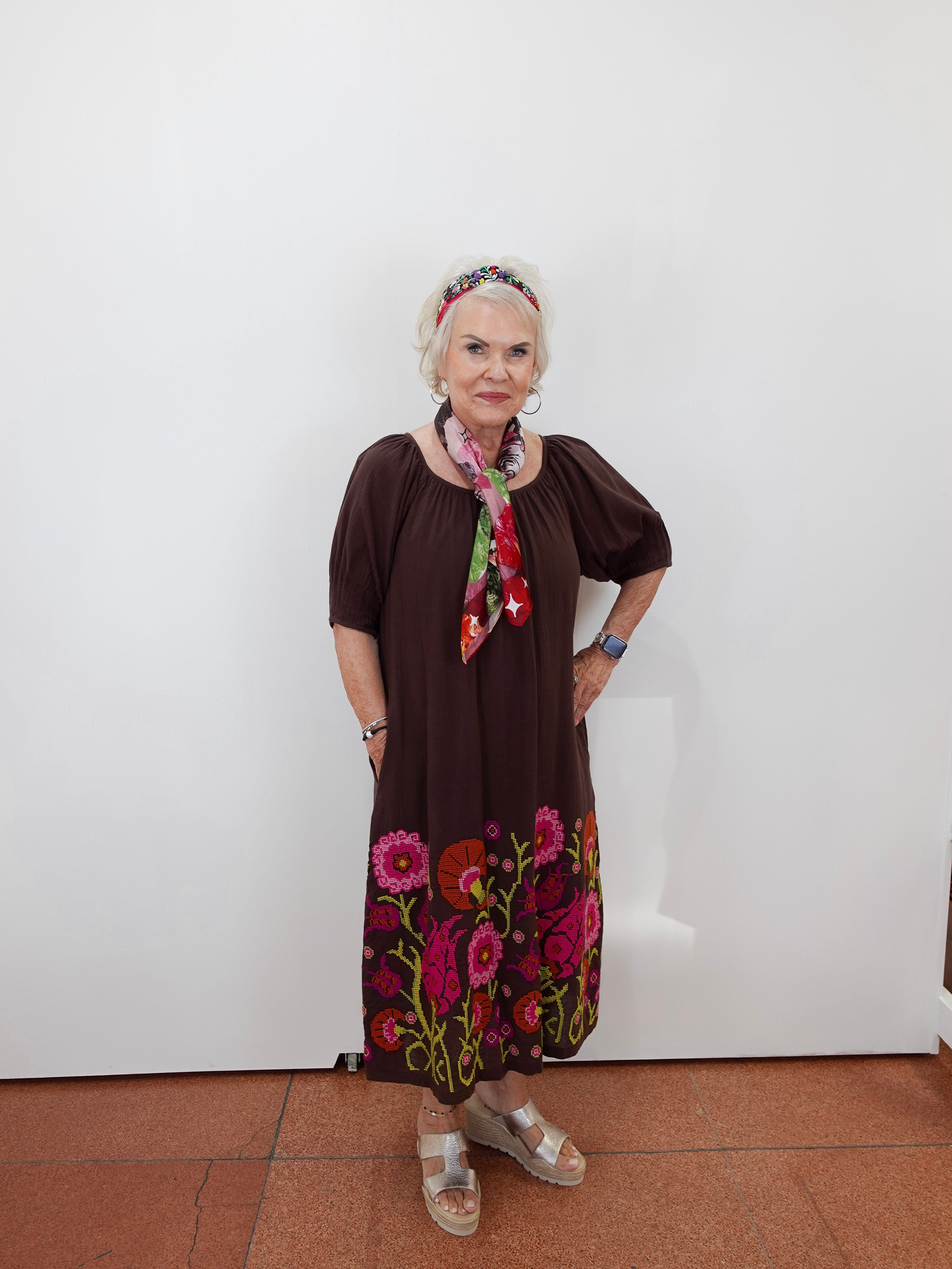 An older woman stands against a plain white wall wearing a dark brown short-sleeve dress with colorful floral embroidery at the hem, a multicolor floral headband, and a matching neck scarf.