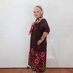 An older woman stands against a plain white wall wearing a dark brown dress with colorful floral embroidery around the hem and a matching patterned headband, paired with sandals and a scarf.