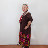 An older woman stands against a plain white wall wearing a dark brown dress with colorful floral embroidery around the hem and a matching patterned headband, paired with sandals and a scarf.