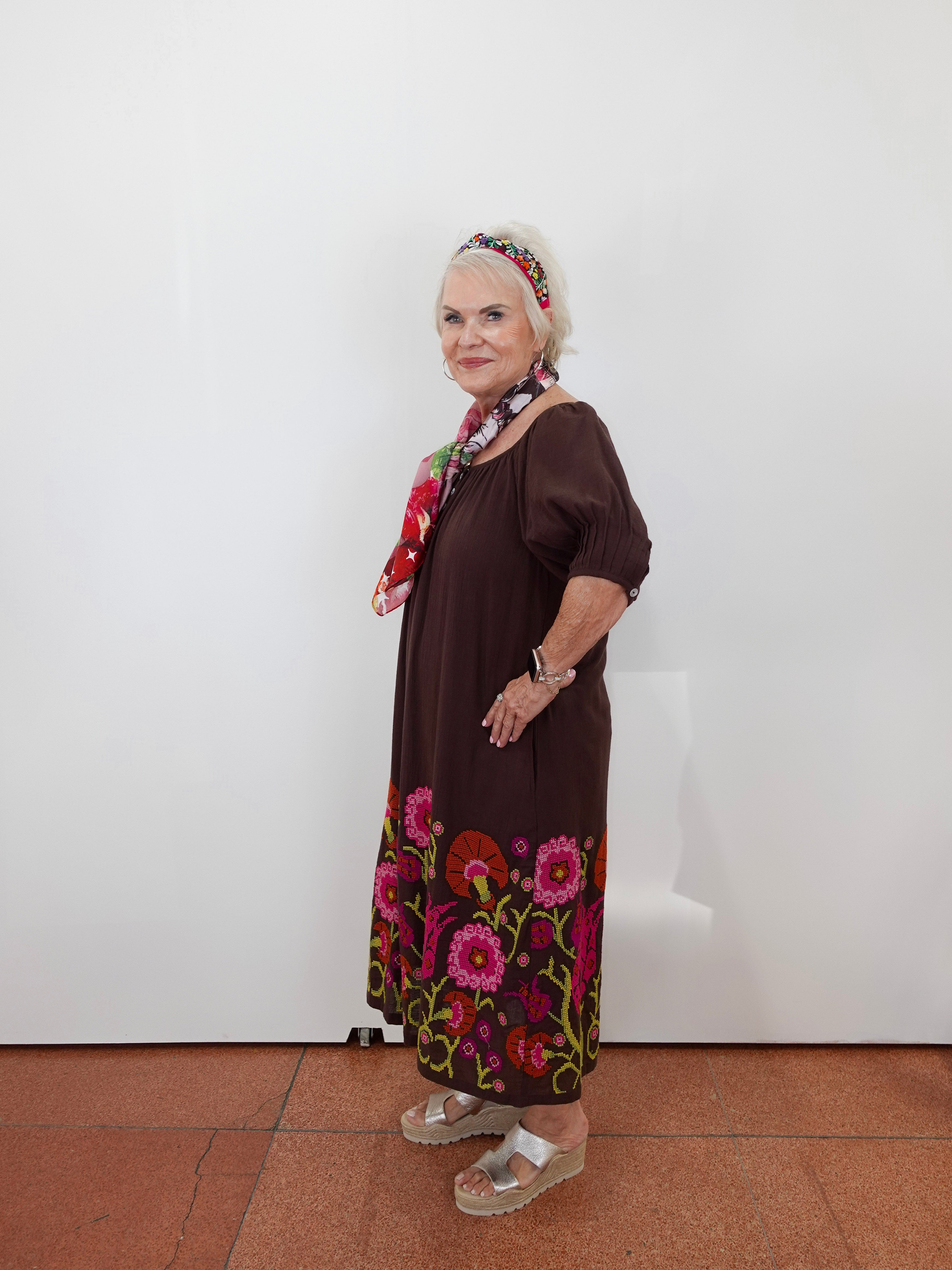 An older woman stands against a plain white wall wearing a dark brown dress with colorful floral embroidery around the hem and a matching patterned headband, paired with sandals and a scarf.