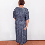 Woman wearing a blue patterned dress standing against a white wall.