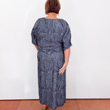 Woman wearing a blue patterned dress standing against a white wall.