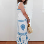 Woman wearing a white dress with blue patterns, holding a beige handbag, standing against a white wall.
