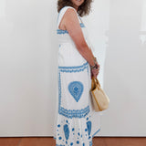 Woman wearing a white dress with blue patterns, holding a beige handbag, standing against a white wall.