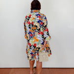 Person wearing a colorful floral dress holding a small handbag against a white wall.