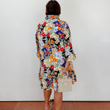 Person wearing a colorful floral dress holding a small handbag against a white wall.