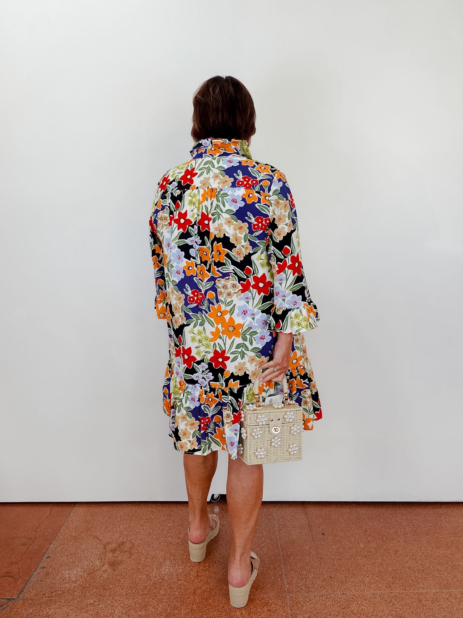 Person wearing a colorful floral dress holding a small handbag against a white wall.