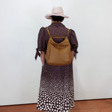 A person viewed from behind wearing a brown and white polka dot midi dress, a light pink wide-brim hat, and carrying a tan leather backpack against a plain white wall.