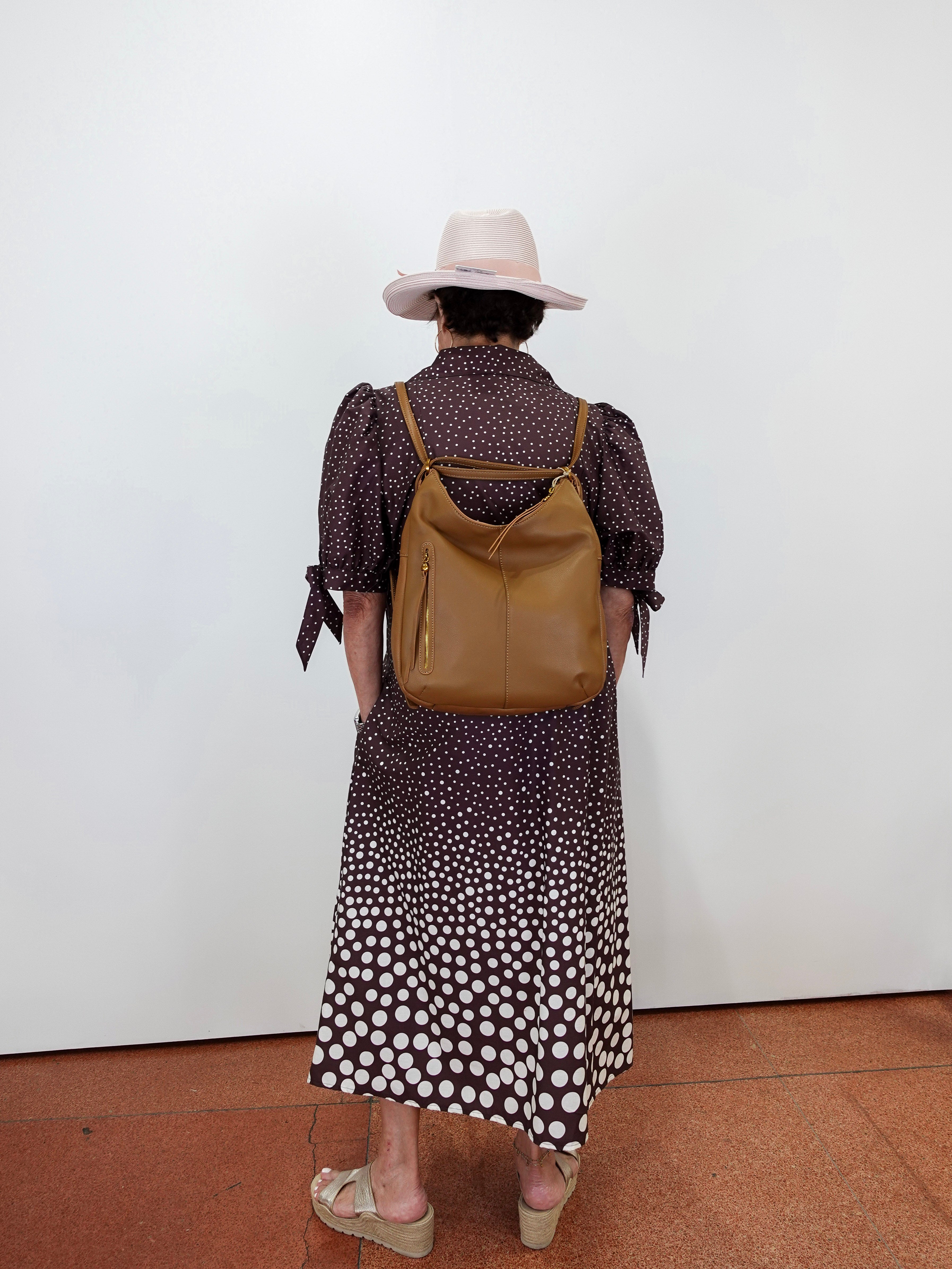A person viewed from behind wearing a brown and white polka dot midi dress, a light pink wide-brim hat, and carrying a tan leather backpack against a plain white wall.