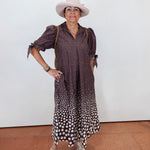 A woman modeling a brown and white polka dot midi dress with puff sleeves, paired with a light beige wide-brim hat, gold bracelets, and beige wedge sandals, standing against a plain white wall.