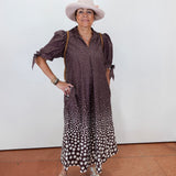 A woman modeling a brown and white polka dot midi dress with puff sleeves, paired with a light beige wide-brim hat, gold bracelets, and beige wedge sandals, standing against a plain white wall.
