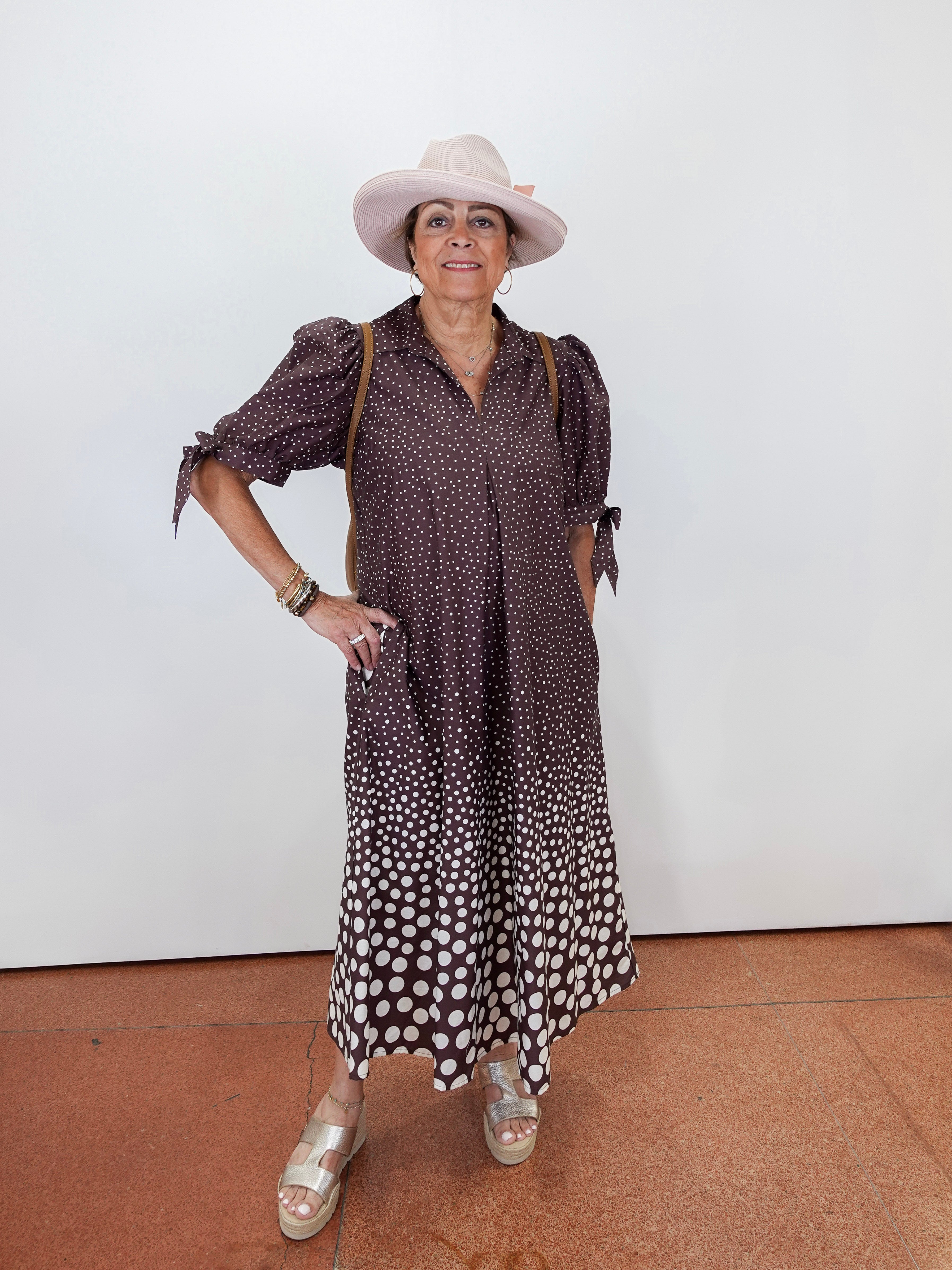 A woman modeling a brown and white polka dot midi dress with puff sleeves, paired with a light beige wide-brim hat, gold bracelets, and beige wedge sandals, standing against a plain white wall.