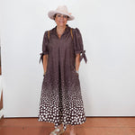 A woman wearing a brown and white polka dot midi dress with tie details on the sleeves, a light pink wide-brim hat, and beige platform sandals, standing against a plain white wall.