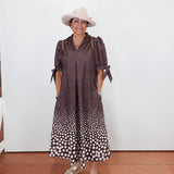 A woman wearing a brown and white polka dot midi dress with tie details on the sleeves, a light pink wide-brim hat, and beige platform sandals, standing against a plain white wall.
