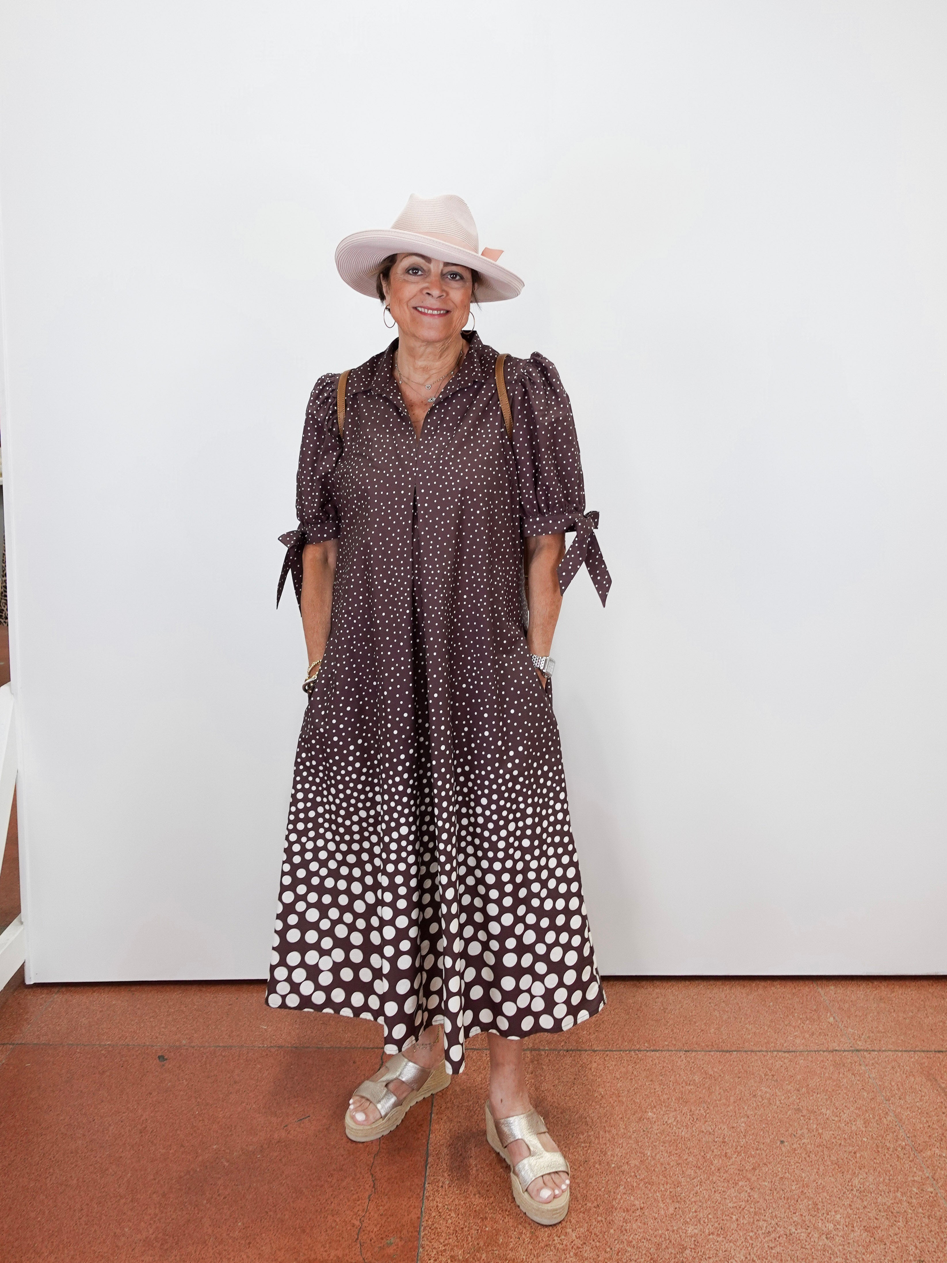 A woman wearing a brown and white polka dot midi dress with tie details on the sleeves, a light pink wide-brim hat, and beige platform sandals, standing against a plain white wall.
