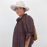 A woman wearing a brown and white polka dot shirt dress, a light pink wide-brim hat, and a tan shoulder bag, posing against a plain white background.
