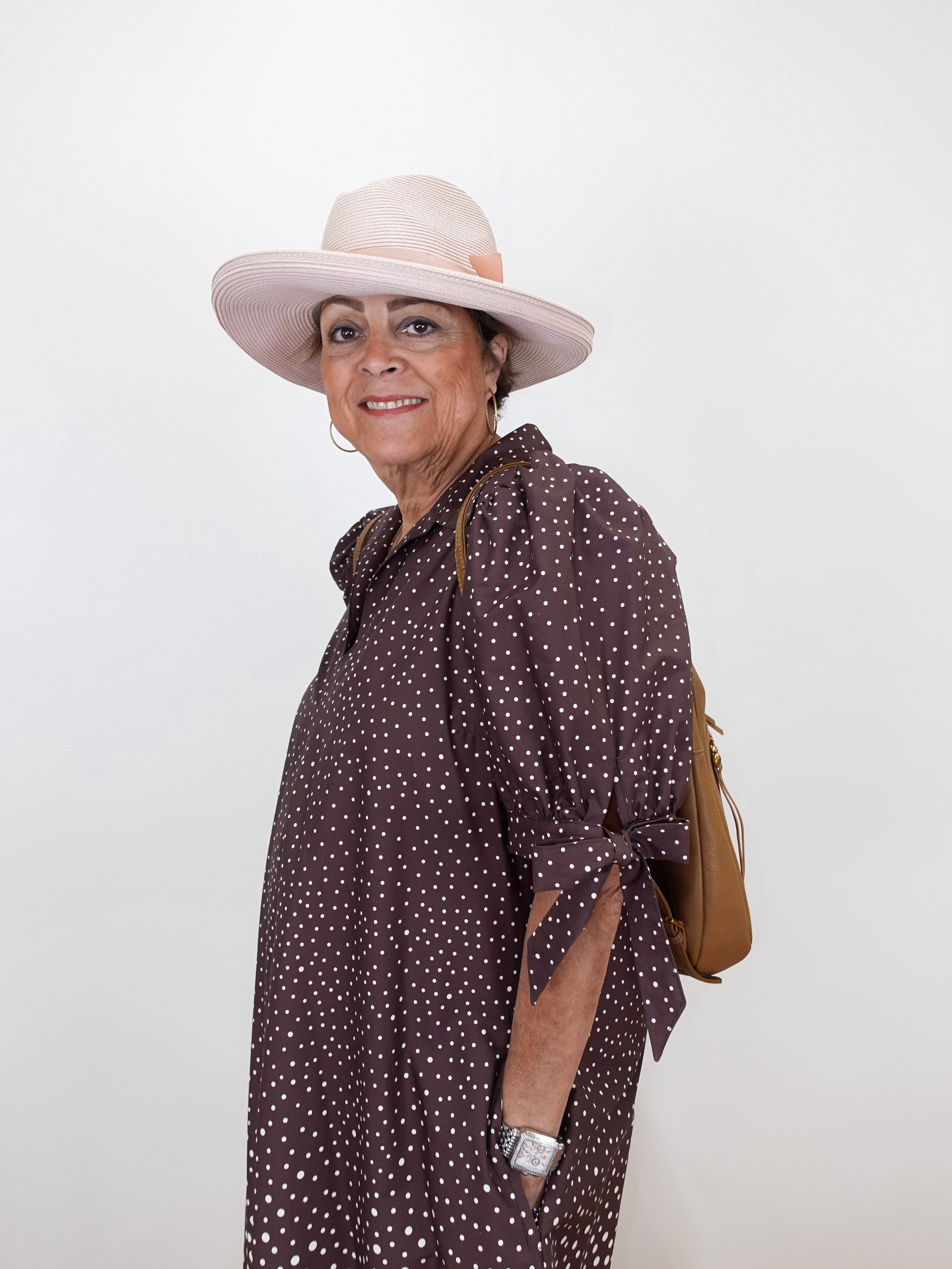 A woman wearing a brown and white polka dot shirt dress, a light pink wide-brim hat, and a tan shoulder bag, posing against a plain white background.