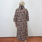 A person viewed from behind wearing a leopard-print coat over a light-colored dress, standing against a plain white wall on a terracotta tiled floor.