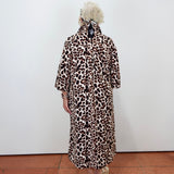 A person viewed from behind wearing a leopard-print coat over a light-colored dress, standing against a plain white wall on a terracotta tiled floor.