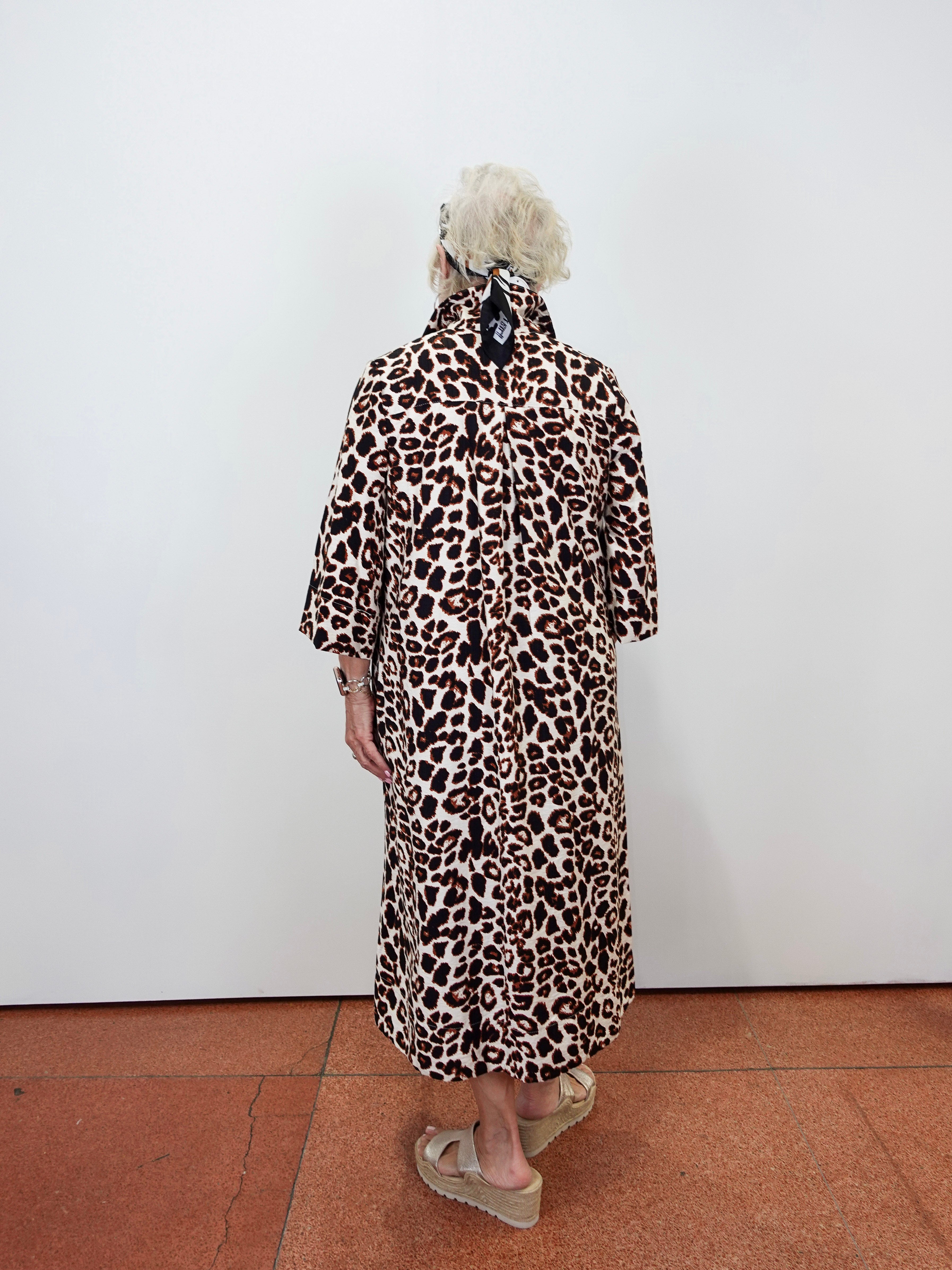 A person viewed from behind wearing a leopard-print coat over a light-colored dress, standing against a plain white wall on a terracotta tiled floor.