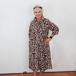 An older woman stands against a plain white wall wearing a long-sleeved leopard-print midi dress with a collar and button front, paired with metallic platform sandals and a headband.