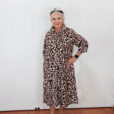 An older woman stands against a plain white wall wearing a long-sleeved leopard-print midi dress with a collar and button front, paired with metallic platform sandals and a headband.