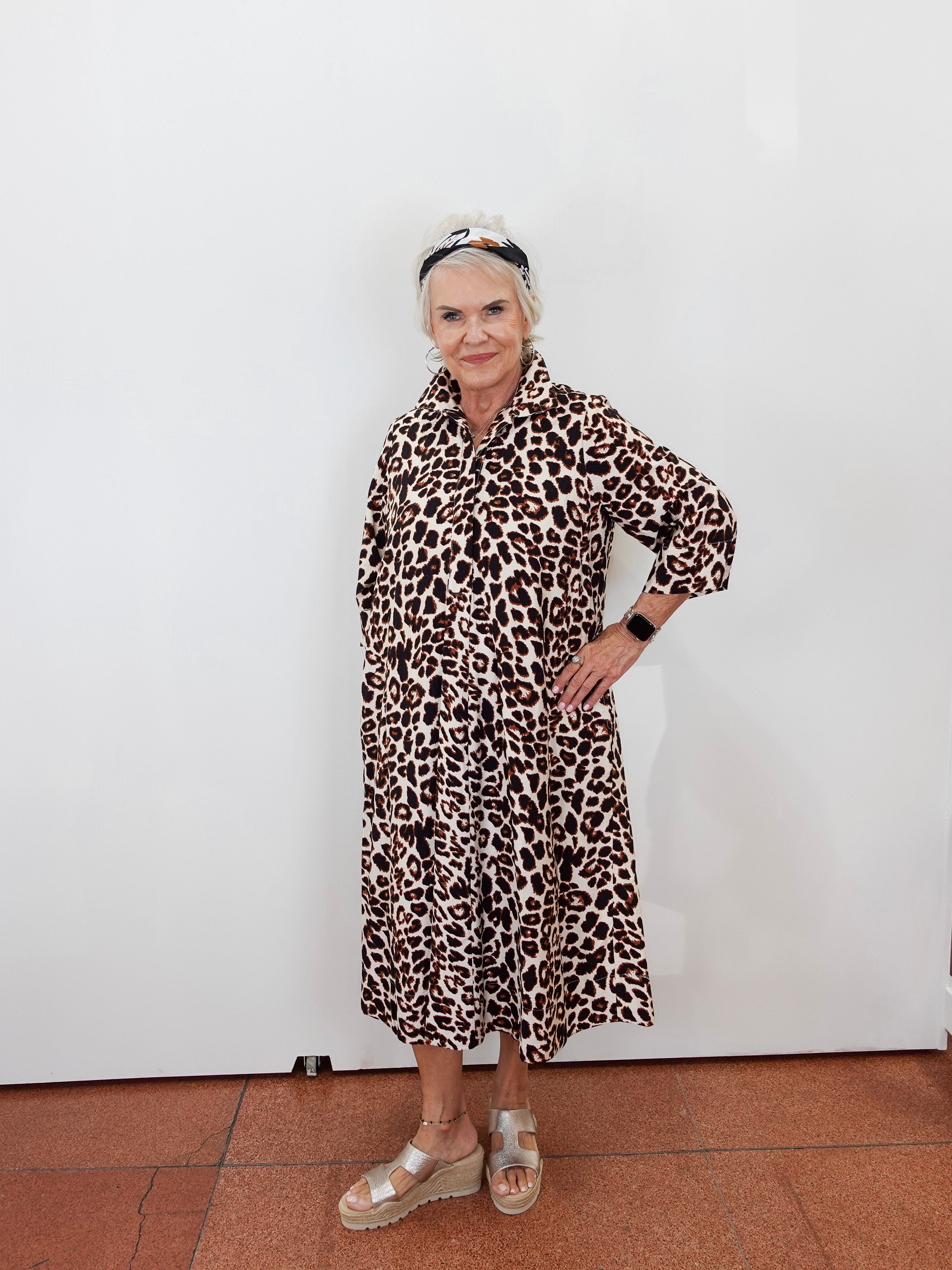 An older woman stands against a plain white wall wearing a long-sleeved leopard-print midi dress with a collar and button front, paired with metallic platform sandals and a headband.