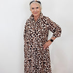 An older woman with short gray hair wearing a leopard print shirt dress, standing against a plain white wall.
