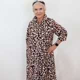 An older woman with short gray hair wearing a leopard print shirt dress, standing against a plain white wall.