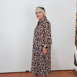 An older woman with blonde hair tied back wears a brown and beige leopard-print midi shirt dress, paired with white platform sandals, standing against a plain white wall.