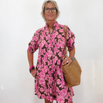 A woman with short blonde hair and glasses models a brown and pink floral print mini dress, holding a tan leather shoulder bag against a plain white wall.